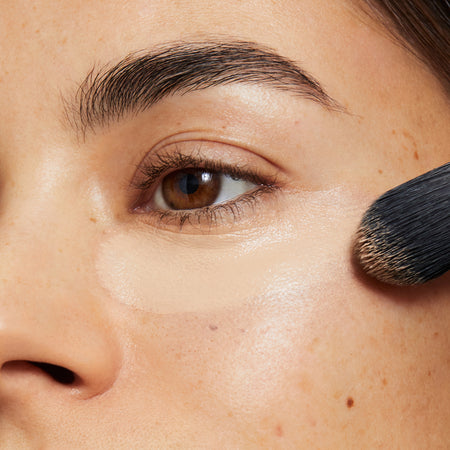 A brunette woman applies creamy beige concealer under her left eye with makeup brush