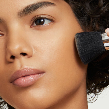 Brunette woman applies powder foundation with makeup brush to her cheek.