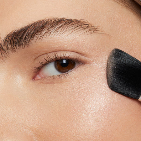 young brunette woman using makeup brush to apply pink blusher powder onto apple of cheek