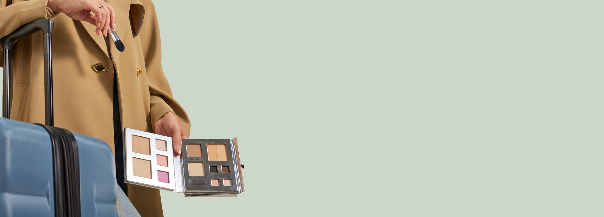 Person holding makeup palettes next to a suitcase on a light green background