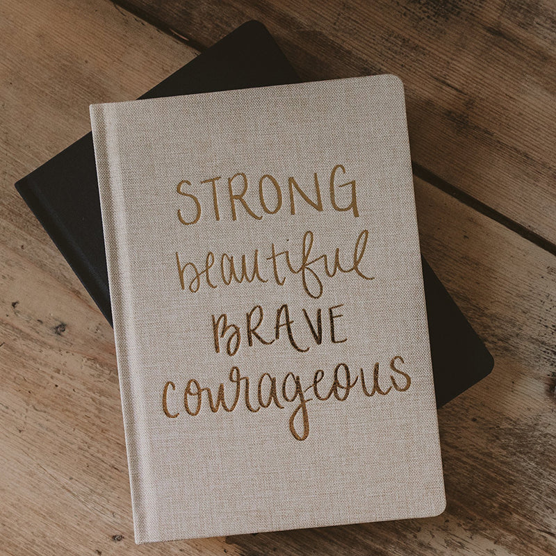  the Strong beautiful brave courageous tan fabric notebook with gold letters displayed on a table