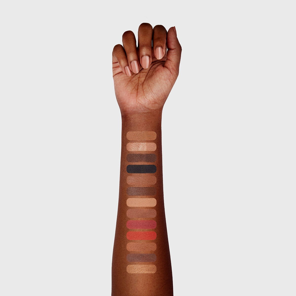 a photo of a dark-skinned woman's arm with 13 swatches showing all 13 cosmetics that are found in the fold out face #5 deep palette