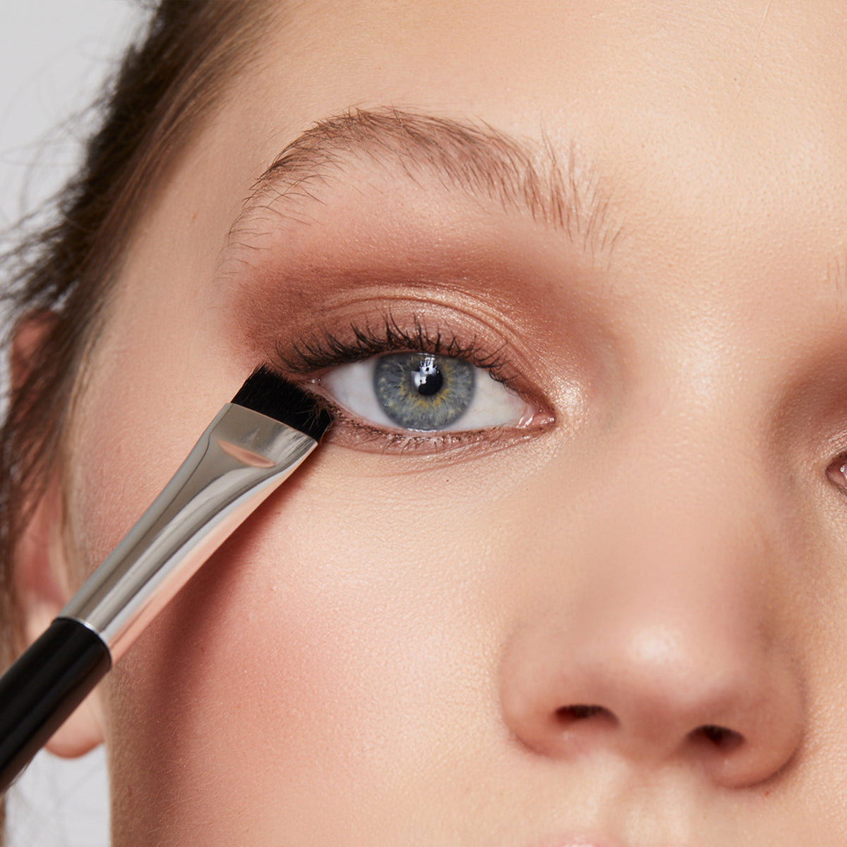 Close up of pretty model with blue eyes holding the angled end of the Corner Brush along her lower lash line to show how bush is used to apply powder eyeliner.