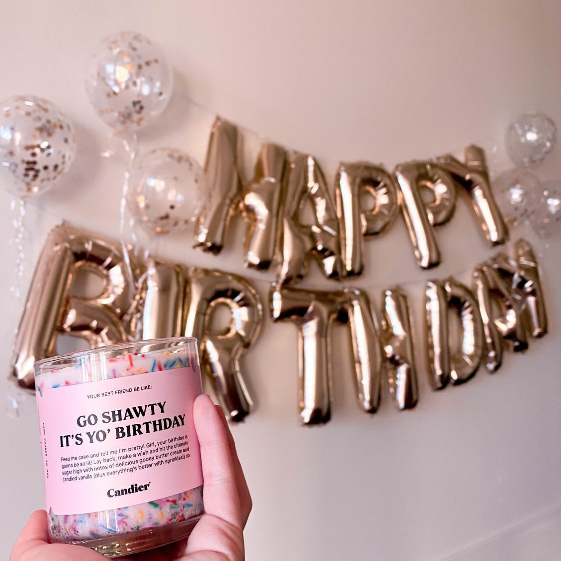 Holding a white birthday candle with sprinkles in clear container in front of a Happy Birthday sign