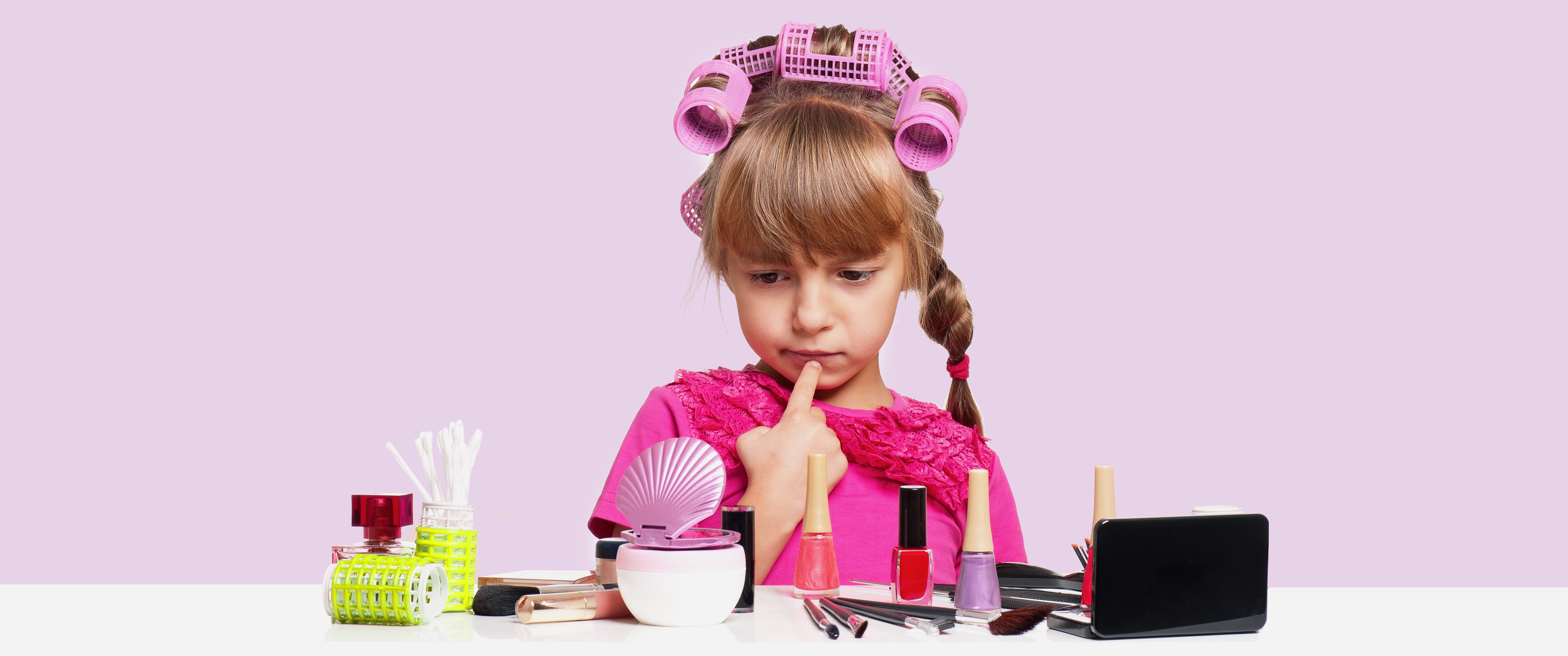 Child in hair curlers looking at different types of makeup products looking confused