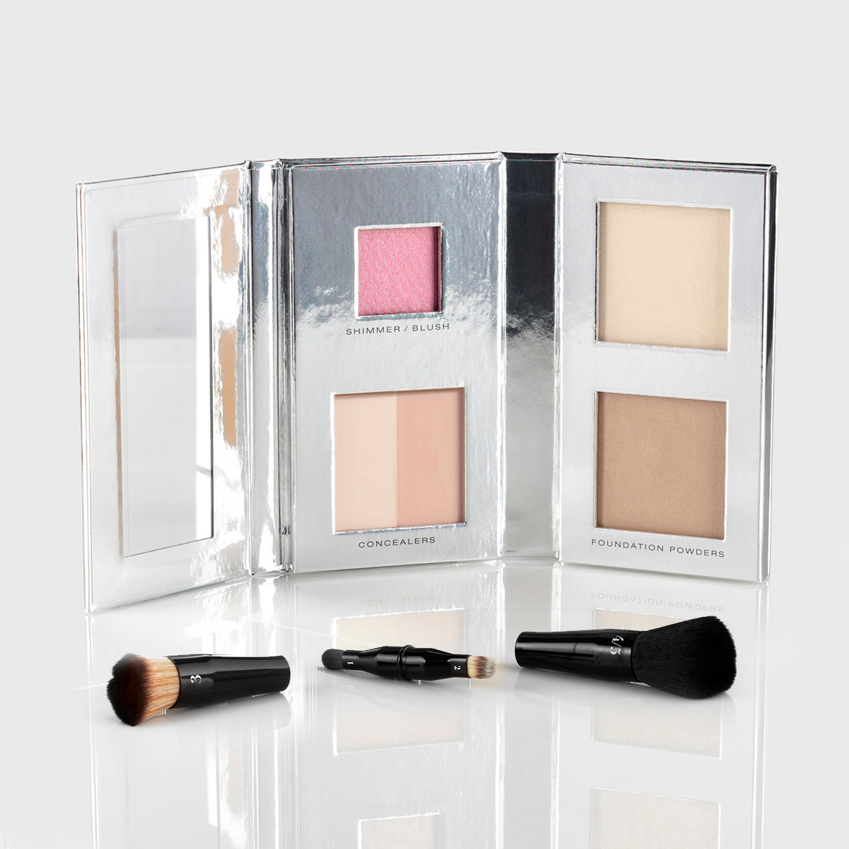a silver makeup palette with mirrored flap and 1 blush, 2 concealers and 2 foundation powers in light shades; a black, travel-sized nesting brush set is splayed out in front of the palette