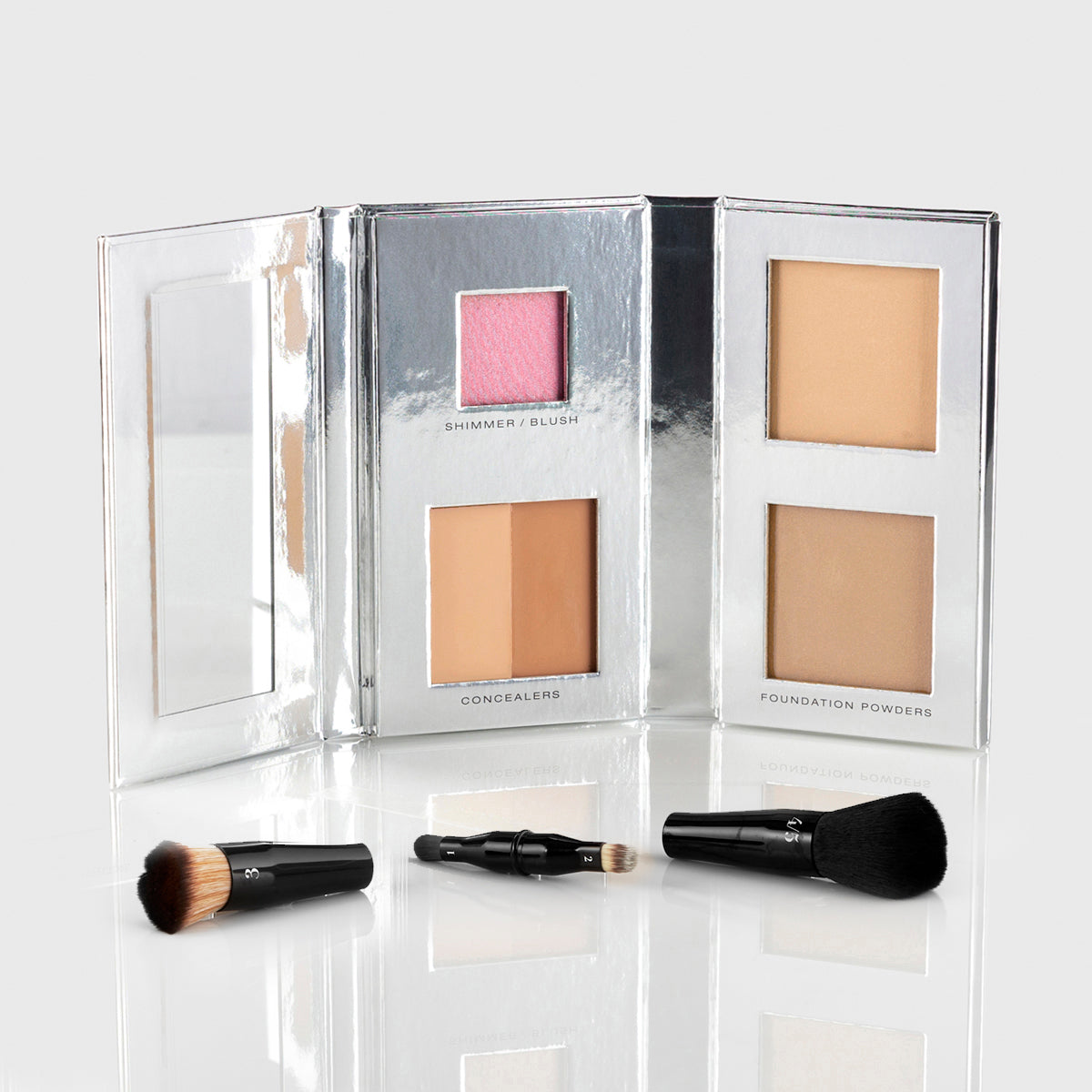 a silver makeup palette with mirrored flap and 1 blush, 2 concealers and 2 foundation powers in medium tan shades; a black, travel-sized nesting brush set is splayed out in front of the palette
