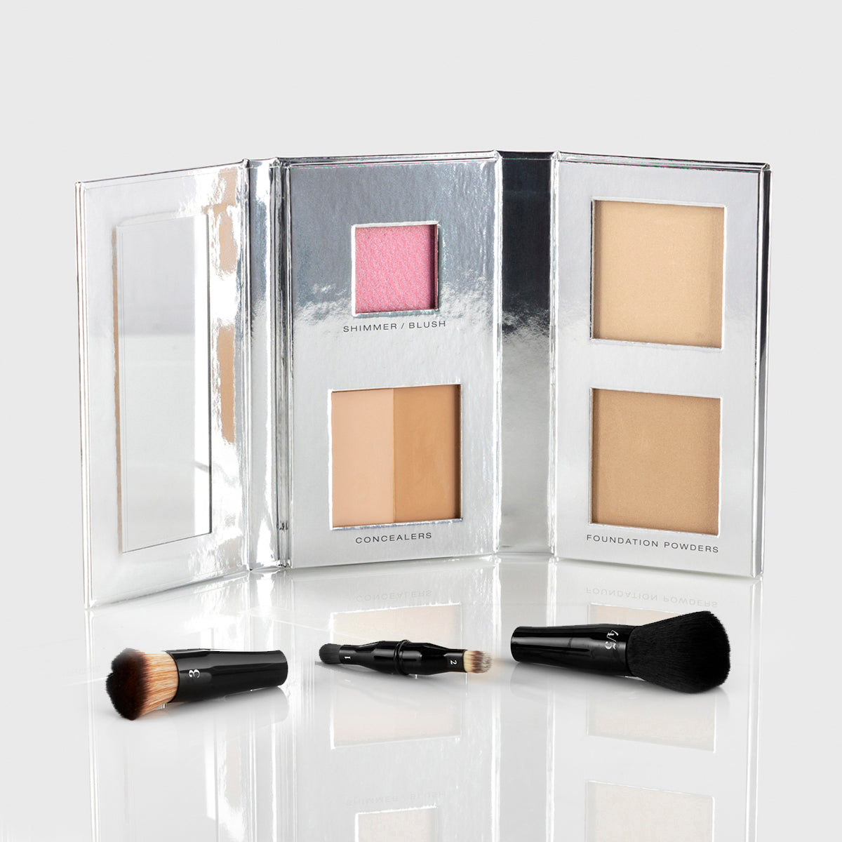 a silver makeup palette with mirrored flap and 1 blush, 2 concealers and 2 foundation powers in medium light shades; a black, travel-sized nesting brush set is splayed out in front of the palette