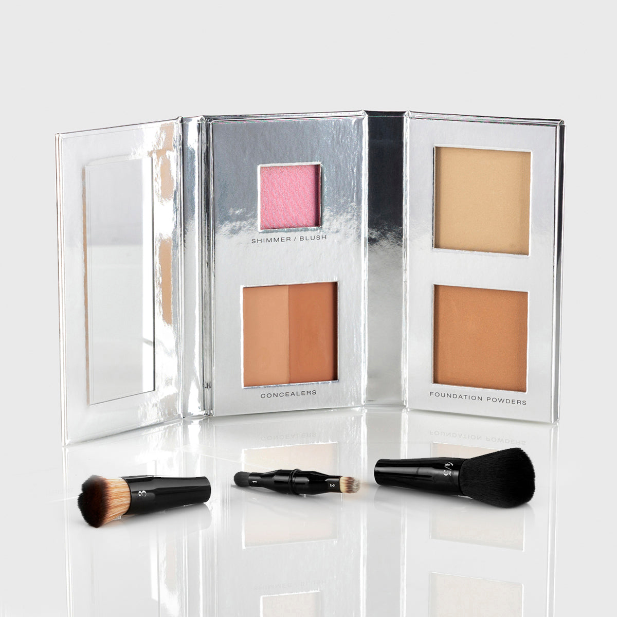 a silver makeup palette with mirrored flap and 1 blush, 2 concealers and 2 foundation powers in medium shades; a black, travel-sized nesting brush set is splayed out in front of the palette