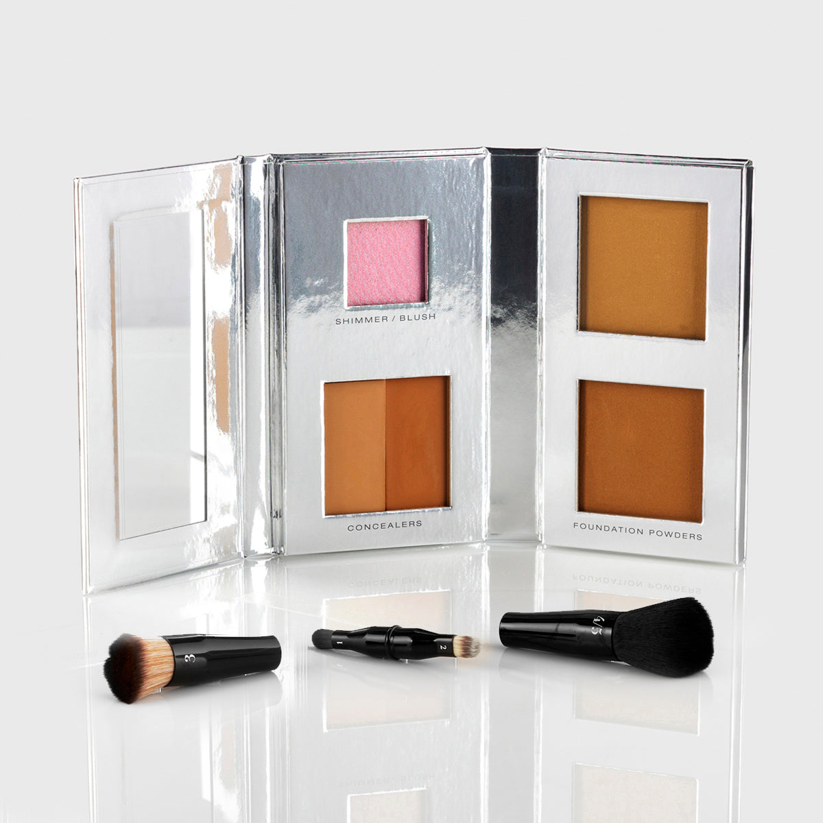 a silver makeup palette with mirrored flap and 1 blush, 2 concealers and 2 foundation powers in medium deep shades; a black, travel-sized nesting brush set is splayed out in front of the palette