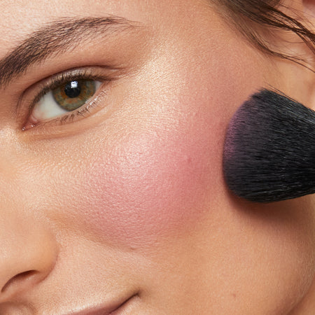 Brunette woman applies pink blush to the apple of her cheek with makeup brush