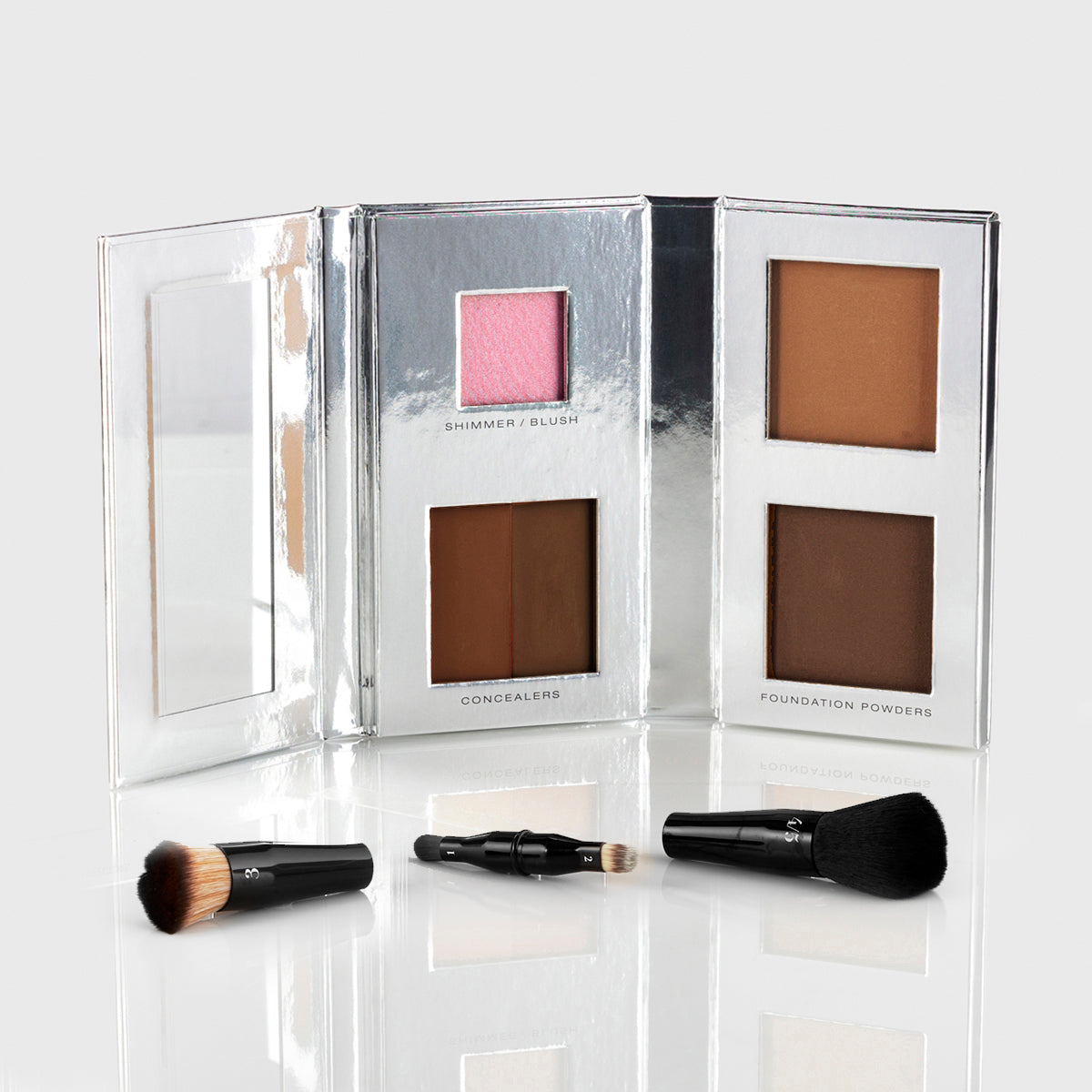 a silver makeup palette with mirrored flap and 1 blush, 2 concealers and 2 foundation powers in deep shades; a black, travel-sized nesting brush set is splayed out in front of the palette