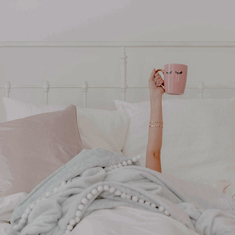 Model holding up the Wakeup to Makeup Coffee mug while laying in bed